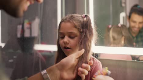 Happy-brunette-man-in-a-green-checkered-shirt-combs-his-little-daughter's-brunette-girl-hairstyle-with-a-ponytail-in-a-pink-dress-in-a-modern-bathroom