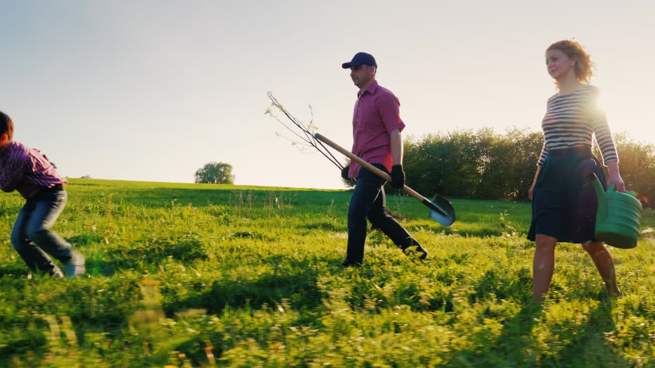 Side view: A family of farmers with a small son go together to plant a tree. Bear the apple tree seedlings, shovel and watering pad. Steadicam shot, side view
