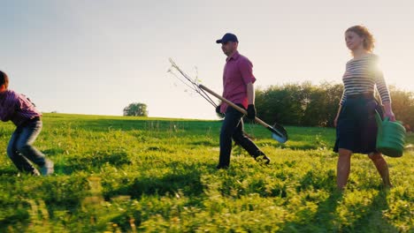 side view: a family of farmers with a small son go together to plant a tree. bear the apple tree seedlings, shovel and watering pad. steadicam shot, side view