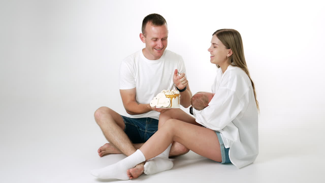 Happy Caucasian couple celebrating the first month anniversary being parents. Woman is holding a newborn in hands. Man lighting a candle on the cake.