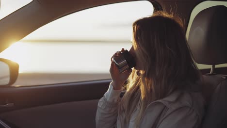 woman inside the car is drinking coffee from the thermos cup, adoring sunrise