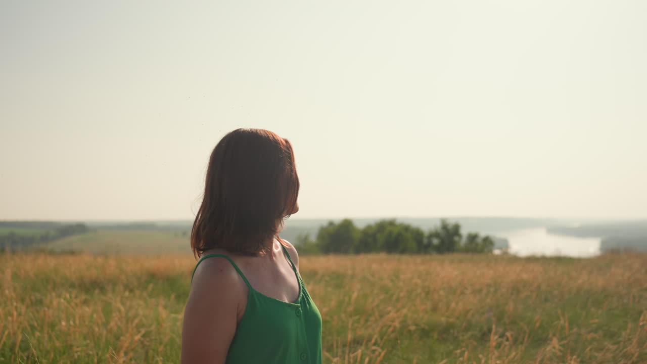 Young woman in green dress stands in golden field with eyes closed, facing horizon and smiling peacefully as warm sunlight bathes landscape in soft glow
