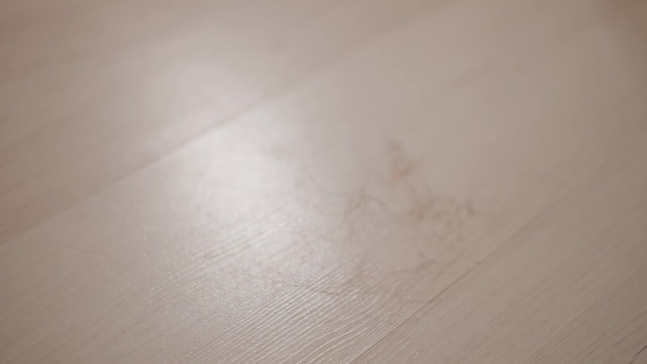Low-angle close-up showing light wood plank floor with warm beige tones, visible grain texture, subtle scuffs, and soft directional light that creates shifting highlight across clean interior space