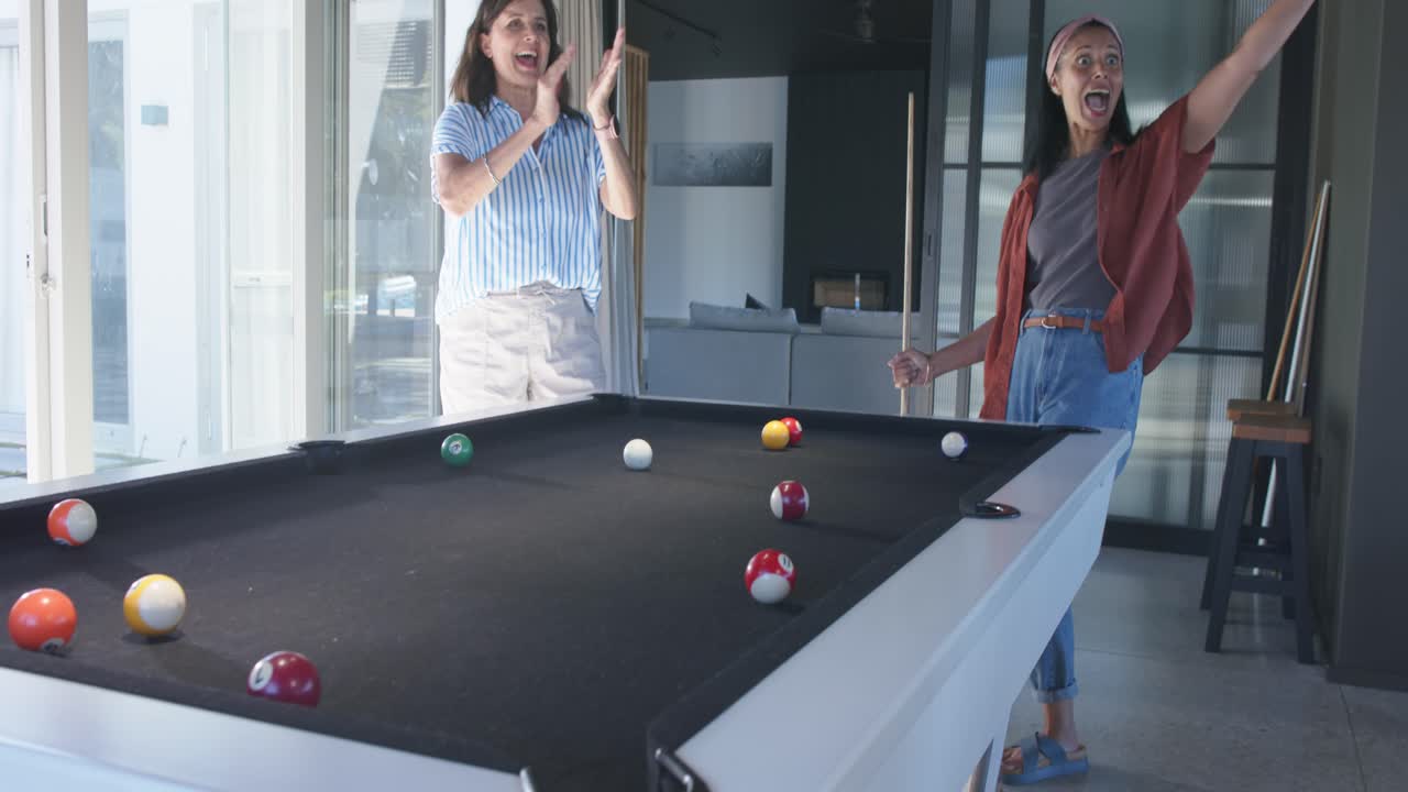Diverse female friends at home pool table watching right woman using bridge, striking balls for fun
