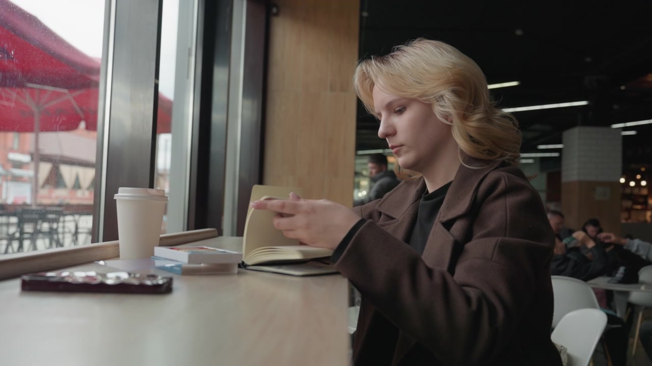 Una donna è seduta a un tavolo in quello che sembra essere un bar o uno spazio pubblico informale, intenta a scrivere su un taccuino aperto. Sul tavolo sono visibili un bicchiere di caffè usa e getta e altri piccoli oggetti