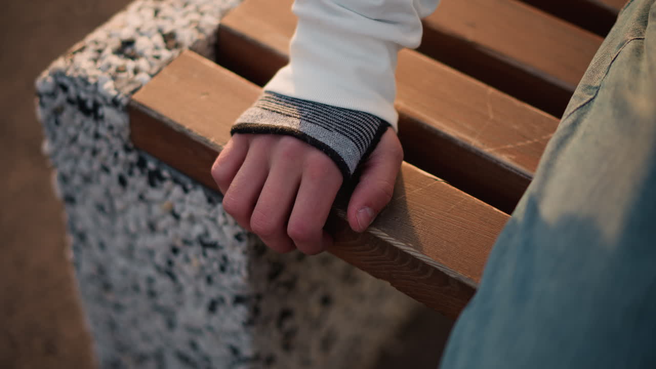 closeup hand on bench wearing fingerless glove at dusk, melancholic outdoor pause with wooden slat and concrete support, denim leg in frame, knit cuff detail, soft golden light, subtle finger
