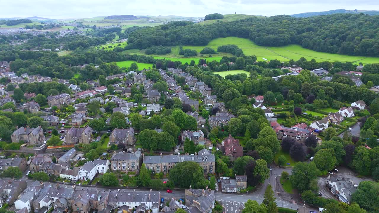Riprese aeree salgono dolcemente sopra un'area residenziale a Buxton, Inghilterra, rivelando file di case, campi verdi lussureggianti e dolci colline in lontananza sotto una luce diurna soffusa.