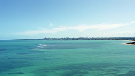 Trucking-right-aerial-wide-shot-of-the-tropical-capital-city-of-Joao-Pessoa,-Paraiba,-Brazil-from-the-famous-Red-Sand-Island-with-turqoise-ocean-water-and-a-large-skyscraper-cityscape-on-a-summer-day