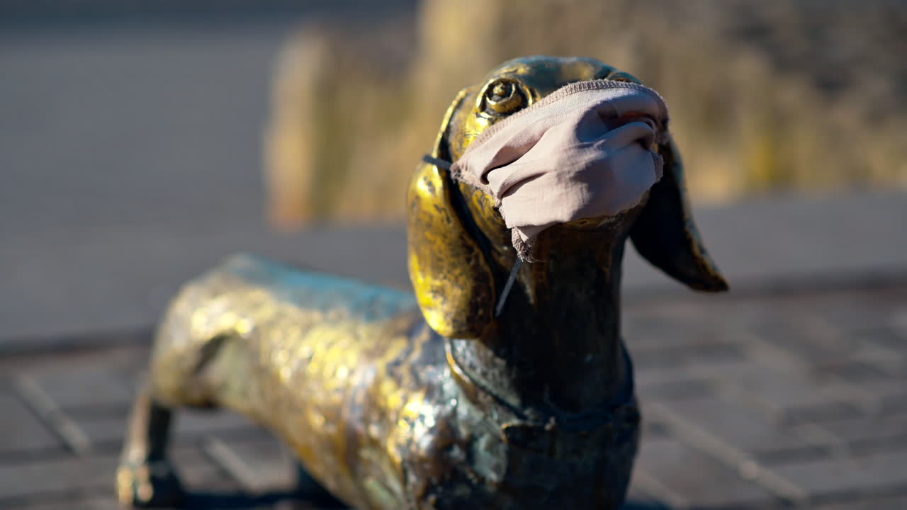 A bronze statue of a dog is seen in a public area, cheerfully displaying a mask. The sunny day highlights the statue's golden details and playful ambiance, attracting passersby