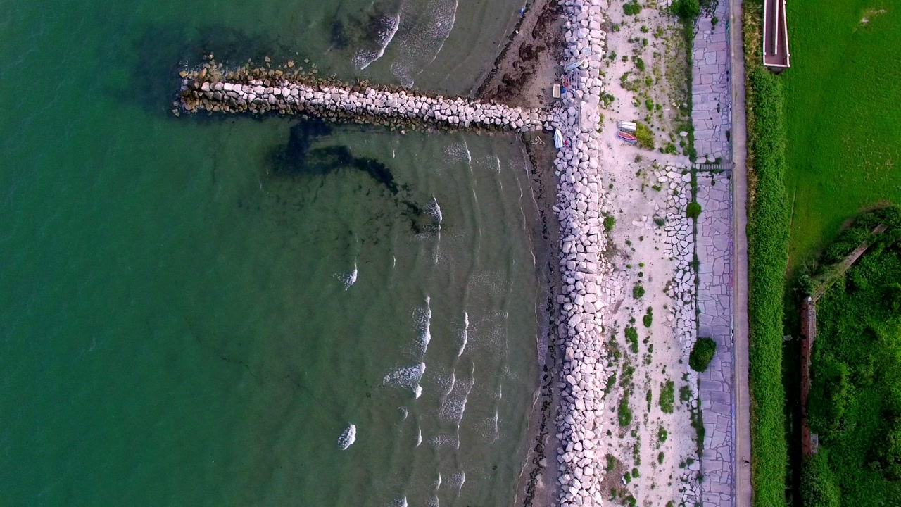 Drone aerial shot looking down over a stone breakwater jetty where shallow turquoise water meets a sandy shore and coastal path at Lido di Venezia.