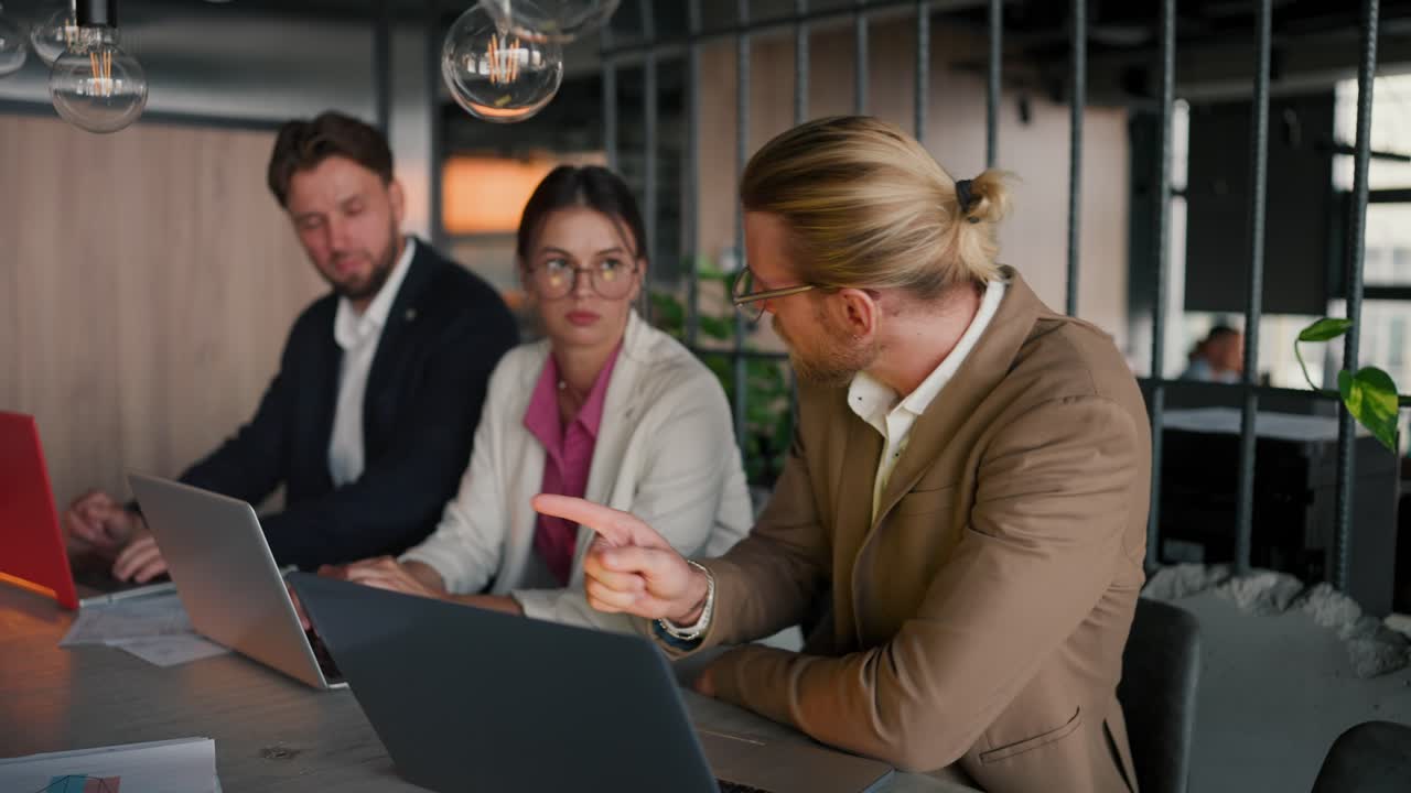 A blond man in glasses with a beard in a light brown suit communicates with his colleagues at the table while caring with a laptop in a modern office