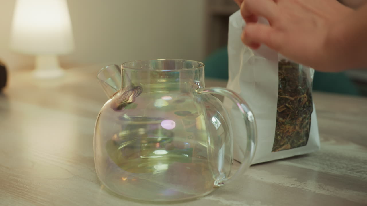 teiera di vetro che versa erbe essiccate in un recipiente trasparente su un tavolo di legno, mani che spargono foglie di lavanda e camomilla, calda luce della lampada e scaffali accoglienti dello studio, primo piano di petali colorati che si depositano, lento