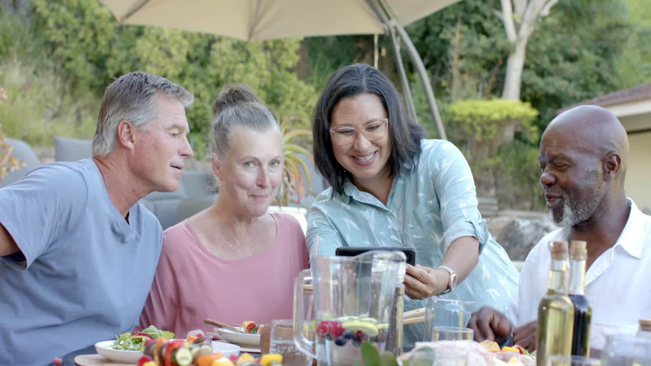 Group of diverse seniors enjoying in garden lunch, smiling and sharing memories on tablet