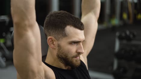 Hombre-En-Forma-Fuerte-Haciendo-Prensas-Con-Mancuernas-Sentado-Durante-El-Entrenamiento-En-Un-Banco