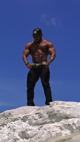 low angle view of the strong fit bodybuilder showing his biceps. handsome athletic man showing his muscles while standing at the sand with blue sky at the background. vertical video