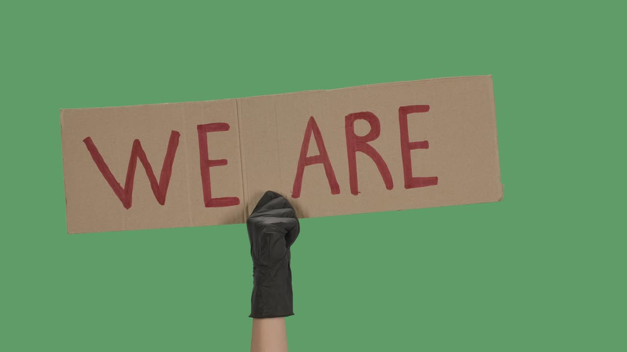 The hand of an unknown person in black gloves raise a cardboard poster WE ARE. Equality and unity concept. Isolated a green screen, chroma key. Close up. Slow motion