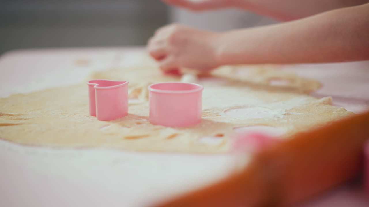 Close up of children hands using pink shape cutters on rolled dough during baking activity, various cutout shapes visible on floured table