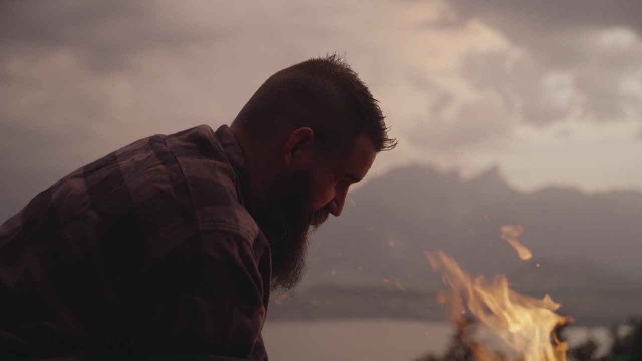 Um homem de camisa de flanela cuida de uma fogueira durante o pôr do sol com montanhas no plano de fundo. Conceito de estilo de vida ao ar livre, aventura, sobrevivência na natureza e conexão com a natureza