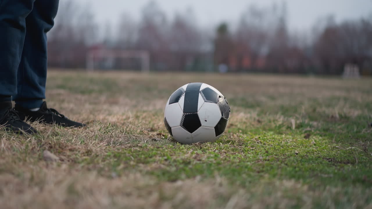 Close-up of a soccer ball on a grassy field, with a person standing nearby and another approaching close to the ball, the background is blurred, showing a football goalpost