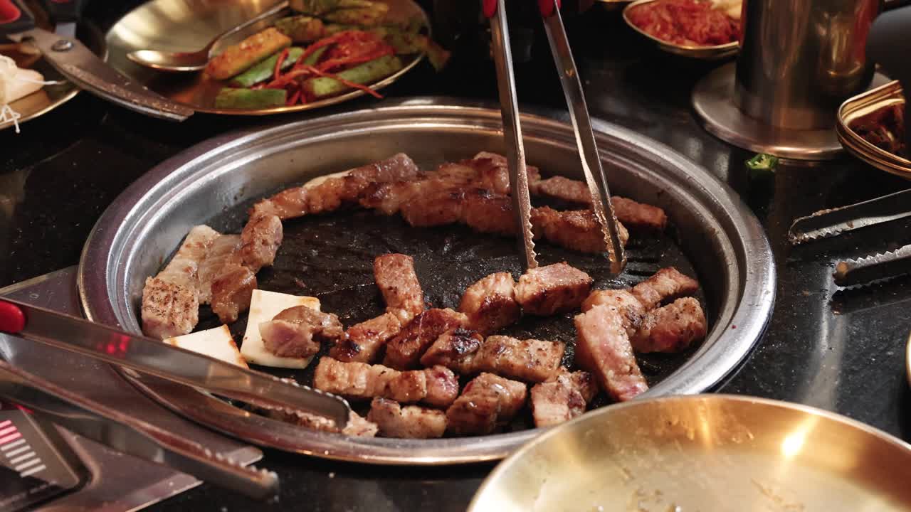 Tongs rotate sizzling pork belly pieces on tabletop grill, surrounded by side dishes, warm lighting