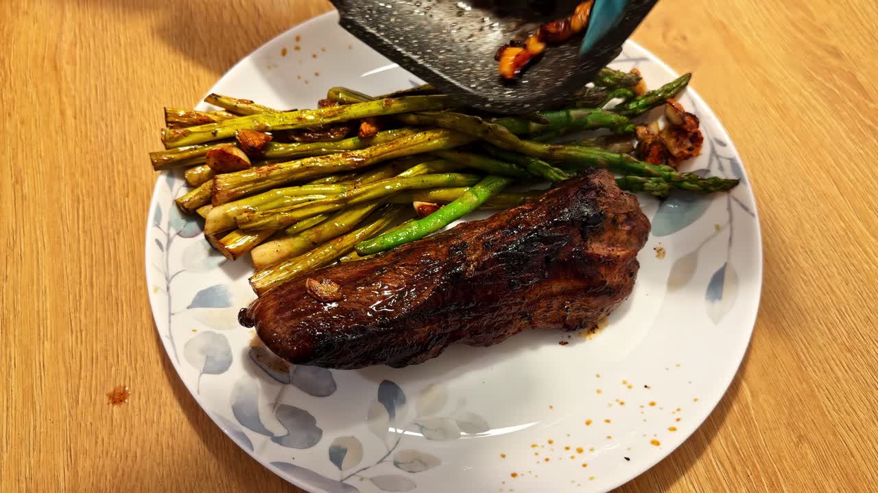 Freshly cooked vegetables slide from a pan onto a plate beside a dark, caramelized meat piece, capturing the moment of serving a homemade, flavorful dish