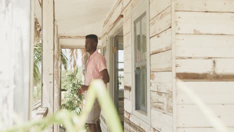 African-american-man-stretching-on-the-porch-of-wooden-beach-house-in-the-morning-sun,-slow-motion
