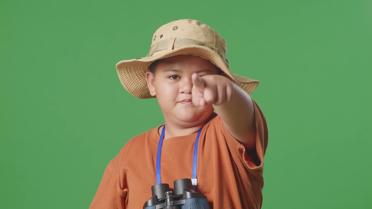 모자와 안경을 쓴 아시아 관광 소년은 보기를 사용하며 손가락을 당신 쪽으로 가리키고 머리를 흔들며 초록색 스크린 배경에 지표손가락을 흔들고 있습니다. 소년 연구자는 제스처를 보여주지 않습니다.