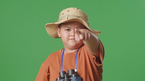 niño turista asiático con sombrero y binoculares usando lupa señalando con el dedo hacia usted, sacudiendo la cabeza agitando el dedo índice en el fondo de la pantalla verde. niño investigador que no muestra ningún gesto, cerrar