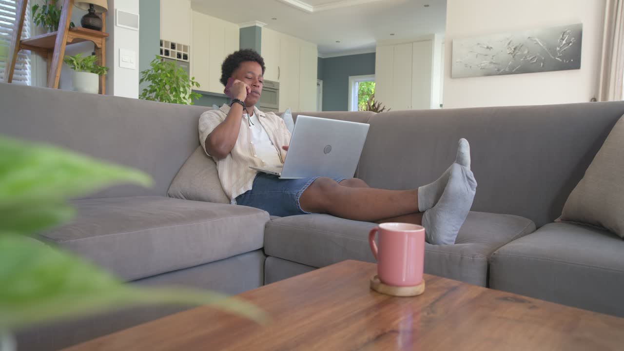 African American man taking call, glancing at laptop, checking red phone, typing, working on sofa