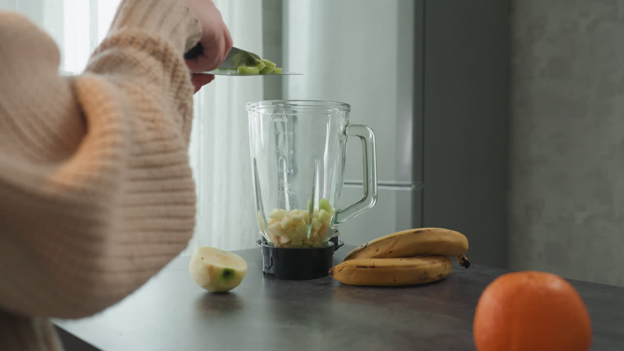 Primo piano di mani che riempiono il frullatore con pezzi di frutta, un coltello stabile aggiunge altri pezzi, mezze banane e un'arancia accanto al barattolo, preparazione di un frullato in stile nutrizionista, un rituale calmo in una cucina domestica minimalista con luce soffusa.