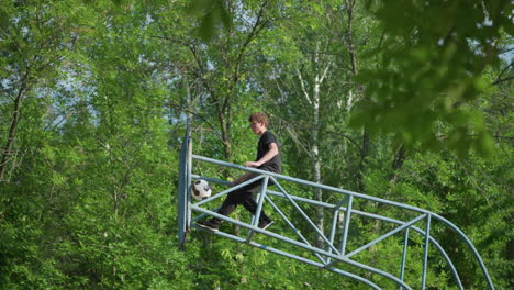 a young boy sits on blue iron outdoor equipment, moving his left hand and leg backward as a ball remains stuck in the framework