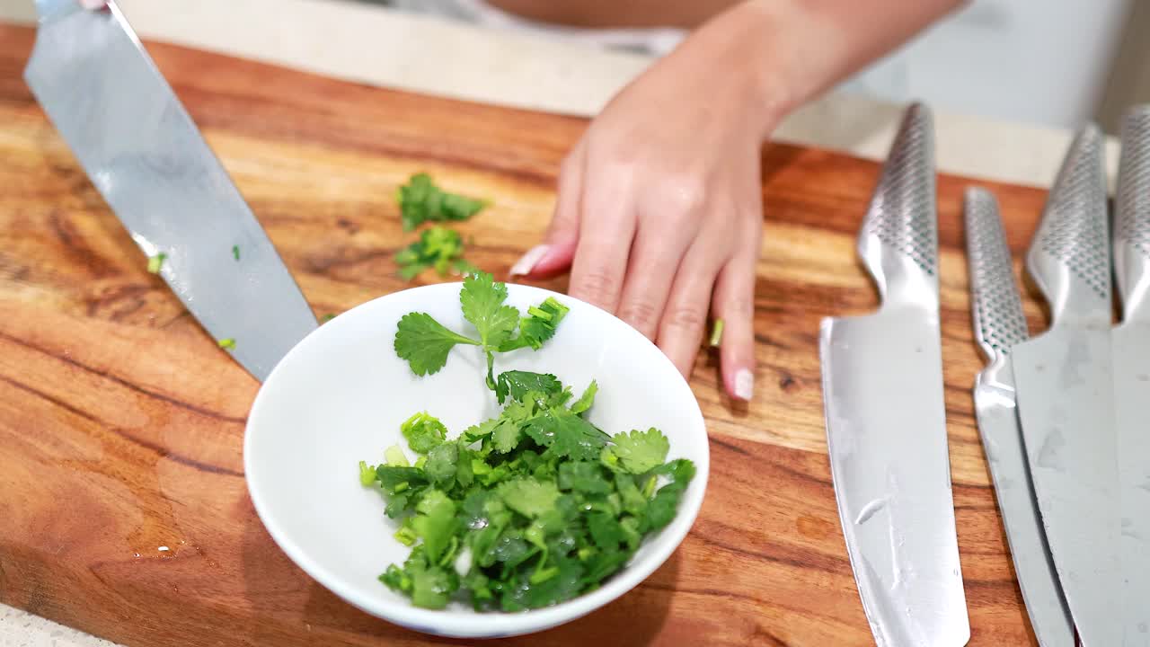 A person chops fresh coriander on a wooden board with precision, using a sharp knife in a well-lit kitchen