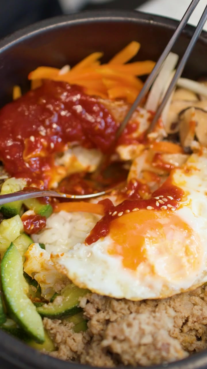 Foto close-up pengadukan bibimbap tradisional Korea dalam mangkuk batu panas menggunakan sumpit.