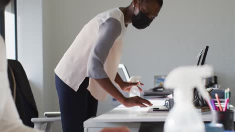 african american businesswoman wearing face mask, sanitizing desk in office