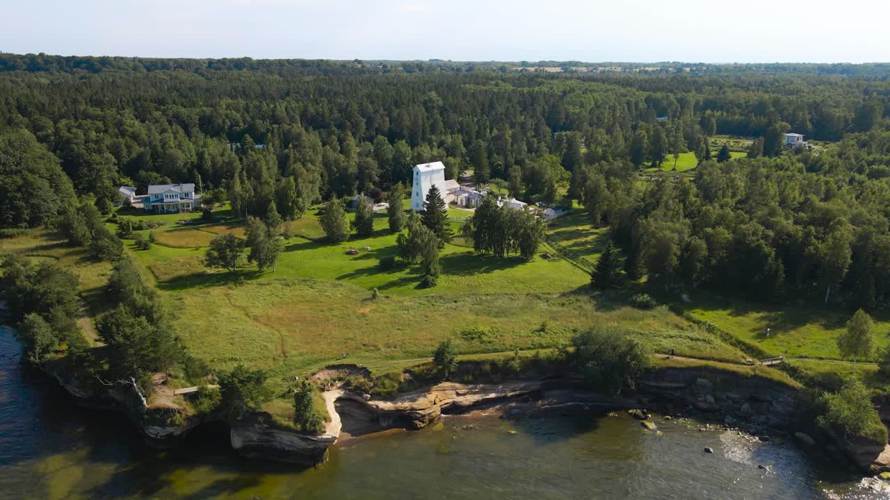 Aerial drone footage flying above and in front of a white colored historic wooden lighthouse in muraste on a sandstone cliff bank beach shoreline or seaside during a summer sunny day. Rocky beach