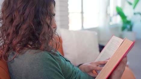 Profile-close-up-shot-of-hispanic-male-with-long-brown-curly-hair-sitting-on-orange-couch-calmly-reading-red-book-in-bright-white-room-infront-of-windows