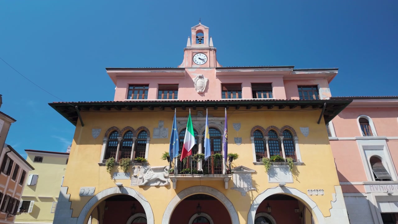 Storico municipio italiano di Muggia-Milje con facciate gialle e rosa, torre dell'orologio, finestre ad arco, balconi, bandiere italiana, europea e locali, e dettagli ornamentali sotto un cielo azzurro limpido