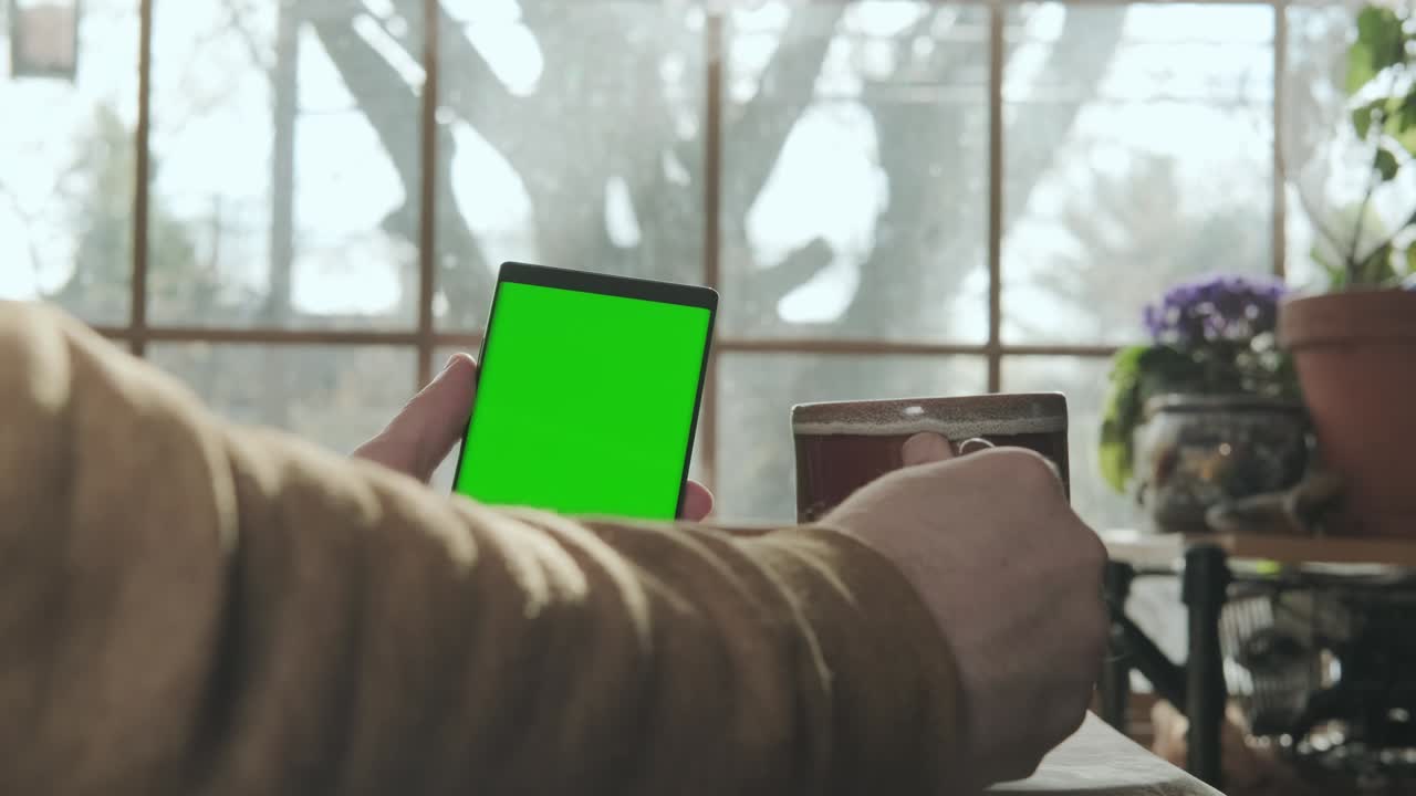 una mano masculina sostiene un teléfono con una pantalla verde de maqueta, y bebe café versión 2