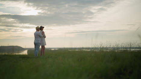 in a peaceful grassland at sunset, a man in a hat, white shirt, and jeans walks towards a woman in a white dress standing on a grassy hill, and embraces her from behind