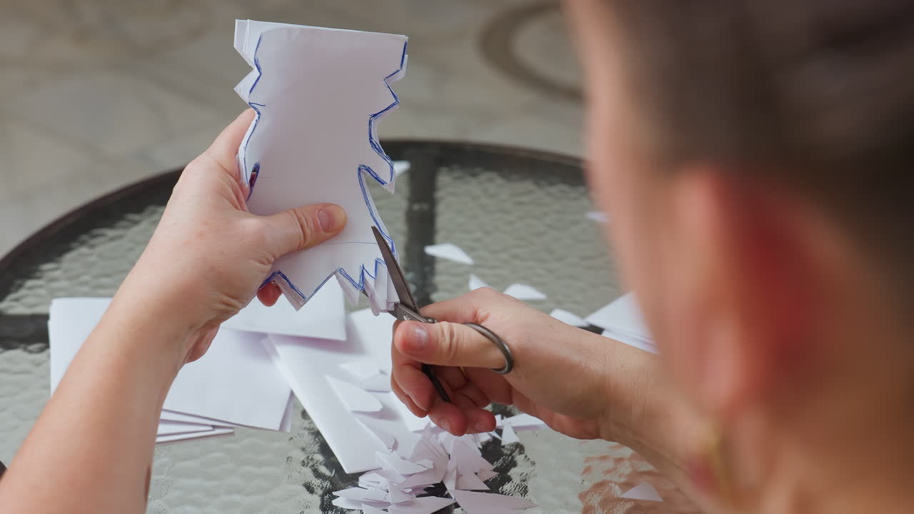 side view of woman carefully cutting white folded paper with blue outlined shapes using scissors on glass table during creative crafting session with small paper pieces scattered around surface