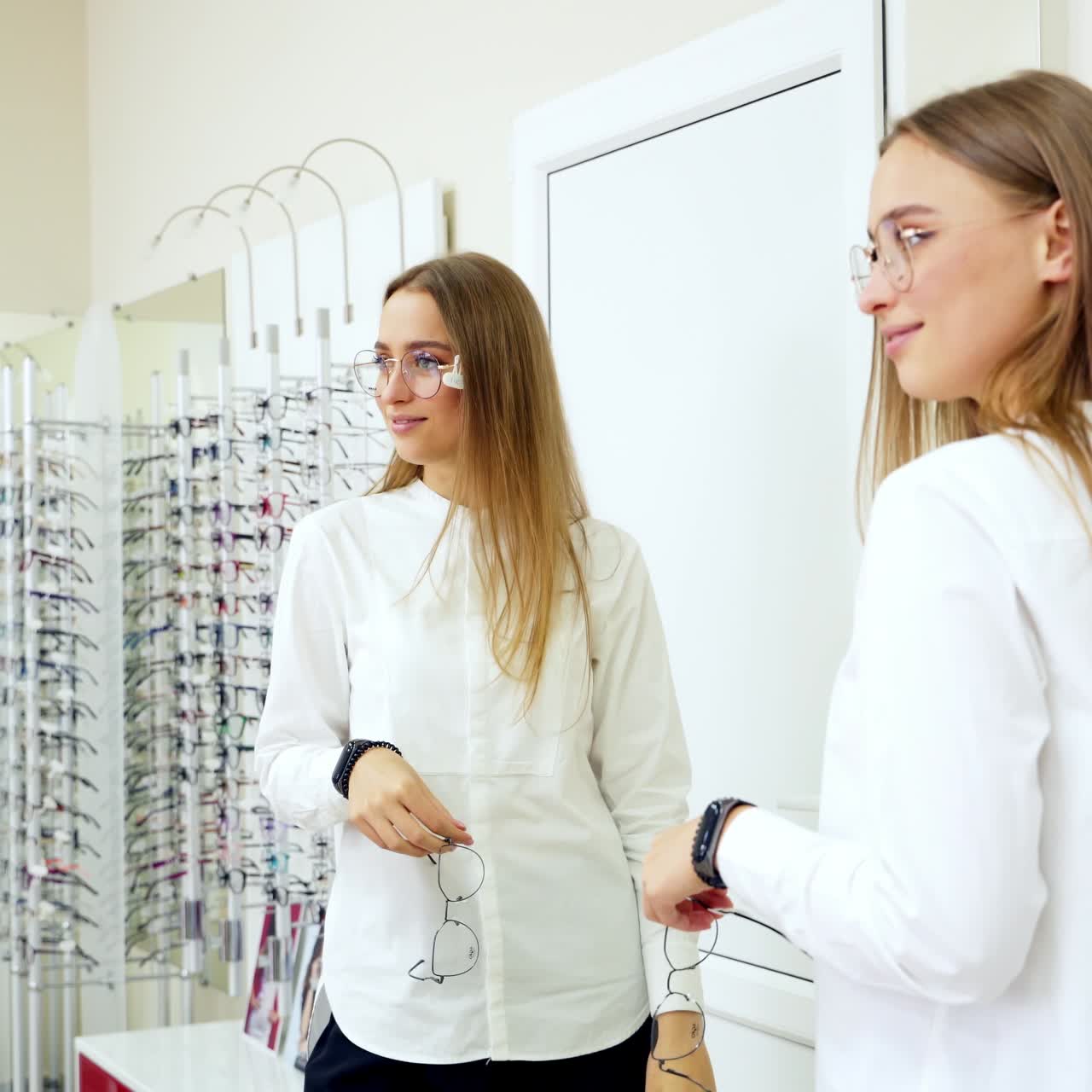 Girl at the optical store. Pretty woman in new spectacles looks at her reflection in the mirror. Eyeglasses choice.