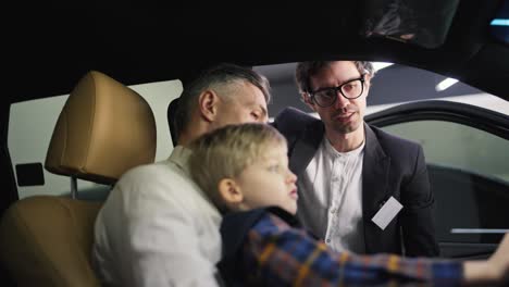 A-confident-middle-aged-man-with-gray-hair-and-a-white-shirt-communicates-with-a-car-dealership-assistant-in-a-business-suit-while-sitting-with-his-little-son-in-the-interior-of-a-modern-car