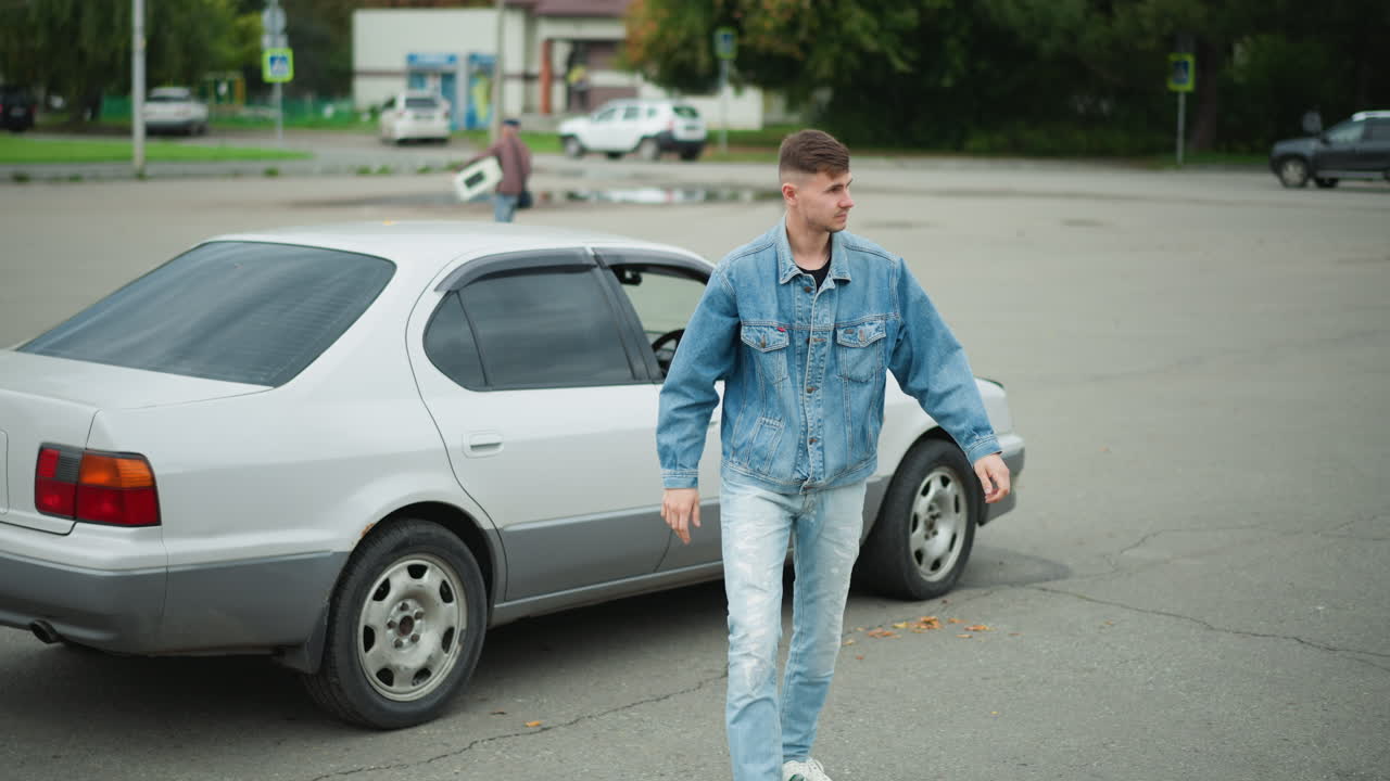 Un giovane uomo bianco scende da una berlina argentata in un parcheggio aperto, indossa una giacca di jeans e un completo di jeans, guarda in lontananza e poi cammina con disinvoltura, stile di vita alla moda con inquadratura cinematografica