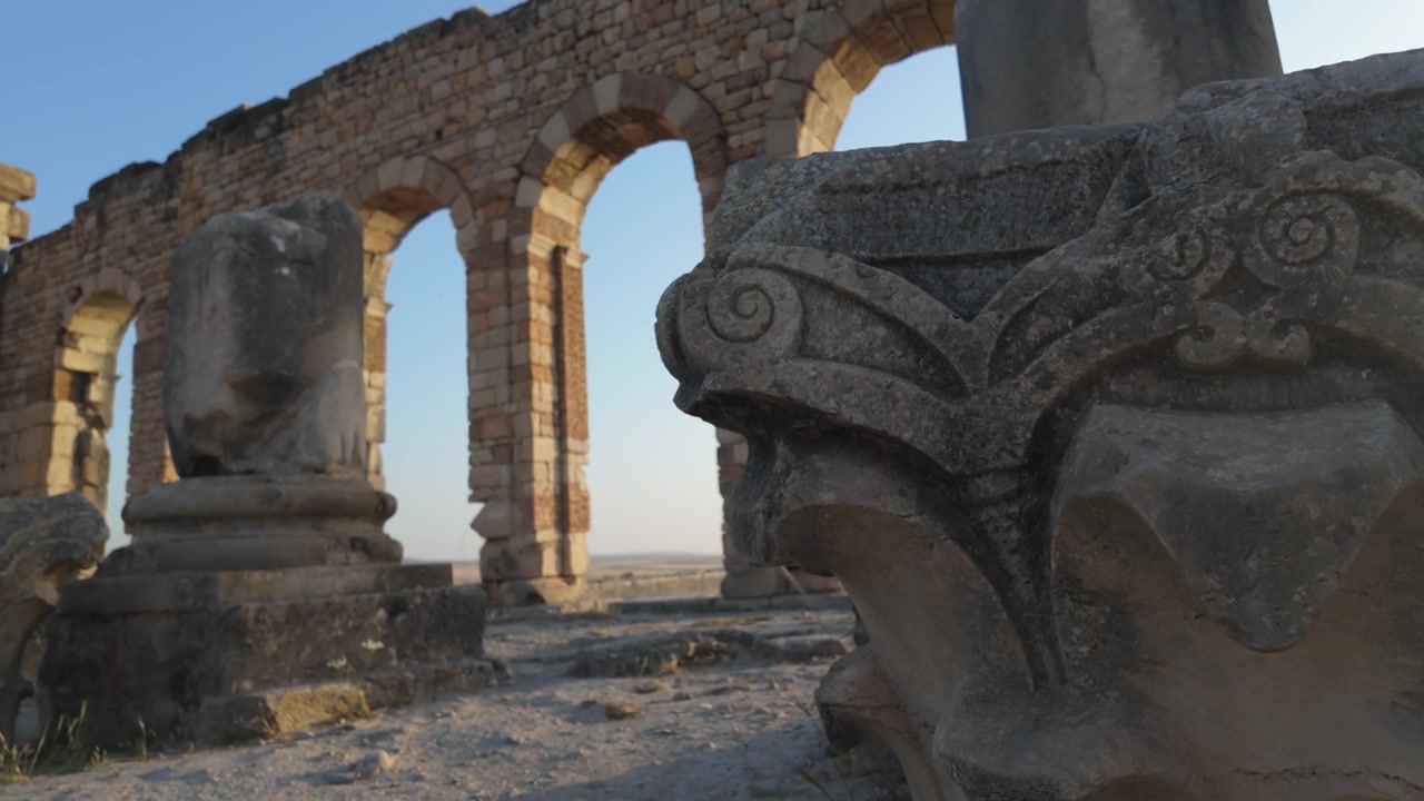 Kinematisk utsikt fra bakkenivå av en korintisk kapitél i forgrunnen med den romerske basilikaens kolonnade bak ved UNESCO-listede arkeologiske stedet Volubilis i Marokko