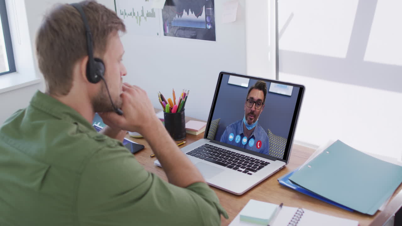 Kaukasisk mand bruger bærbar computer og telefonheadset på videoopkald med en mandlig kollega.
