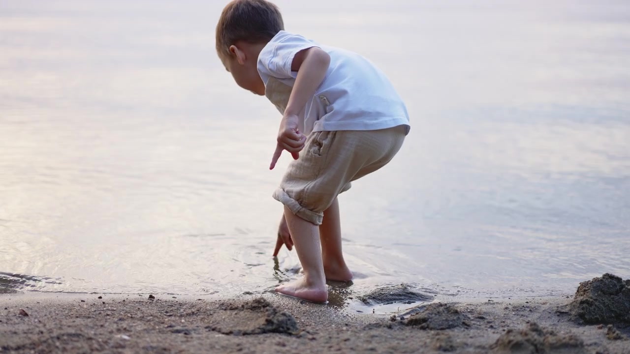 Bambino piccolo che si sporge in avanti al bordo di una riva sabbiosa. Inquadratura dal basso di un bambino che si protende verso l'acqua con sabbia bagnata e leggere increspature sotto.