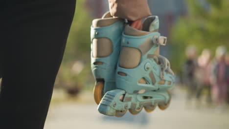 close-up of a person holding a pair of rollerblades, wearing black trousers, walking through a park with a blurred background of trees and people, capturing a casual and leisurely moment