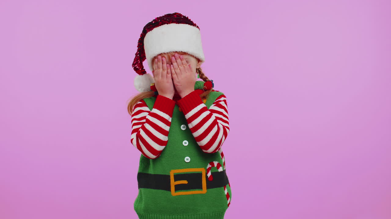 Malá holčička batole v kostýmu vánočního skřítka Santy dovádí, zakrývá si oči rukou a vykukuje skrz.