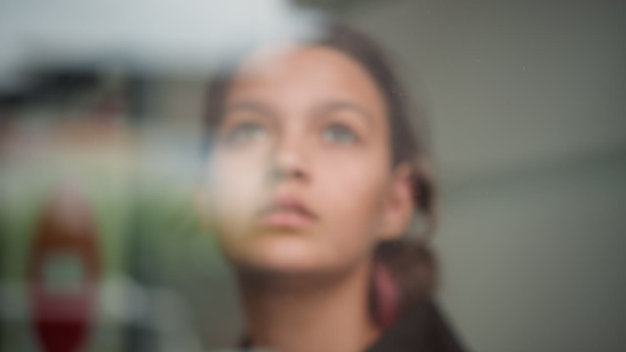 Olhar pensativo na chuva. Adolescente olha através da janela molhada pela chuva. Jovem mulher olha suavemente através do vidro chuvoso. Adolescente melancólica espiando pela janela molhada com paisagem borrada