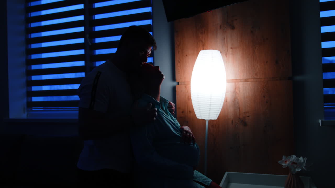 Expecting couple in the dark ward with night lamp. Man stands behind his wife holding her by the shoulders and kissing.